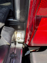 Close-up of a vehicle's rear quarter panel showing the black plastic latch assembly, silver metal bracket, and red taillight assembly.